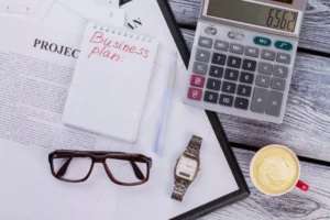 flat lay view of calculator, watch, glasses, and note pad with business plan written on it for starting a small business