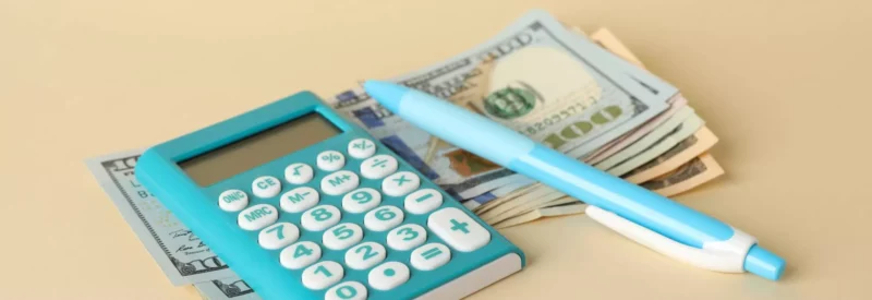 A calculator being used as a business expense tracker on top of a stack of money and next to a pen