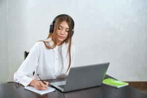 woman in front of laptop on an adult education class writing in a notepad with headphones on 