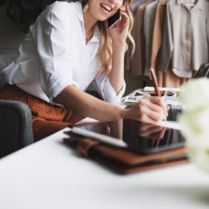 a woman at her fashion studio writing down a brand narrative while she speaks on the phone with tools on her desk 