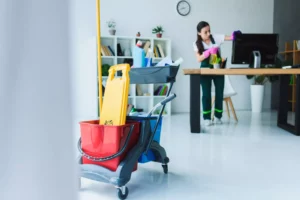 a woman working at her cleaning business, wiping down a monitor in an office with her cleaning supplies next to her. 