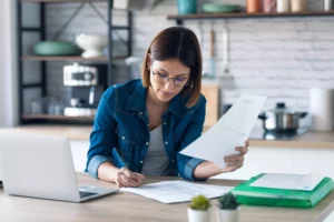 woman sitting at her kitchen counter going over documents in front of her laptop to learn how to make an invoice