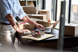 a merchant standing in front of their desk looking at sezzle reviews on a laptop with a tape gun and shipping boxes around them