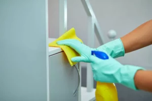 someone starting a cleaning business, wiping a shelf down while wearing gloves and holding a spray and rag