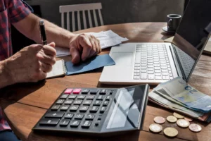 A man writing on a table with a laptop, calculator, and money to figure out what is an ach payment