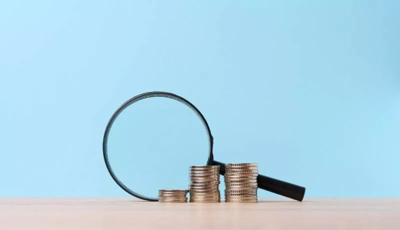 A magnifying glass next to a stack of venmo seller fees coins against a baby blue background.