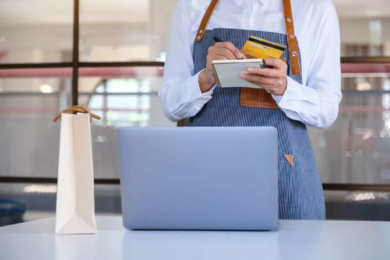 A merchant writing stripe vs clover on a notepad in front of a laptop.