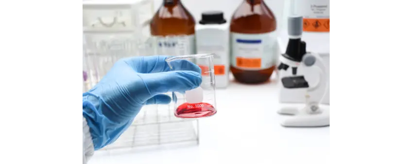 Blue gloved hand holding a carafe with red liquid and bottles of banned ingredients in the background.