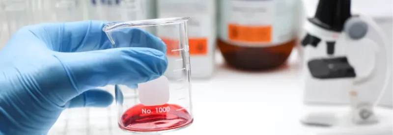 A blue-gloved hand holding a carafe with red liquid and bottles of banned ingredients in the background.