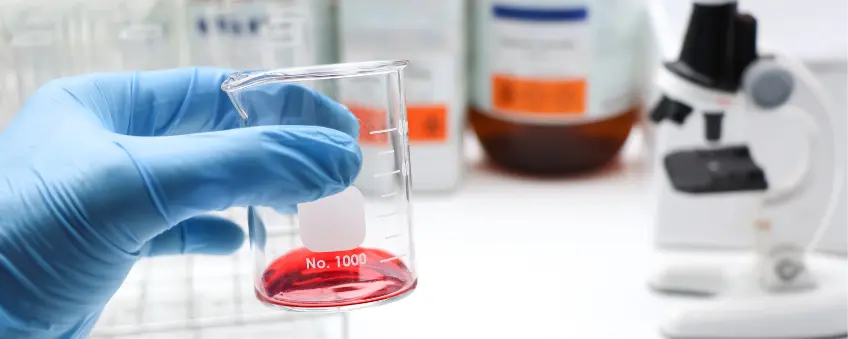 A blue-gloved hand holding a carafe with red liquid and bottles of banned ingredients in the background.