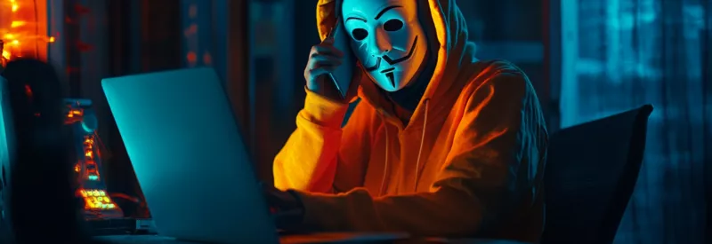 Guy wearing an orange hoodie and a white mask calling someone over the phone while looking at his laptop.